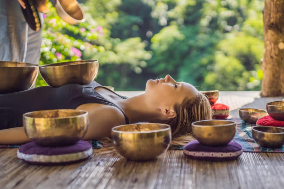 In the Spa, a young gorgeous woman is practicing massage therapy with singing bowls beside a waterfall. 
