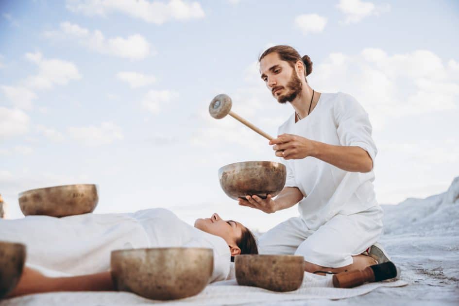 A guy uses Tibetan singing bowls to treat a girl who is resting on the ground in the desert, surrounded by copper bowls.